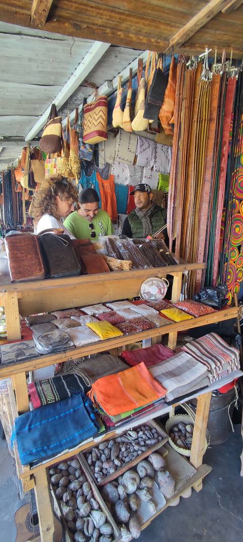       Travelers examine packets of spices at a wooden market stall while the vendor observes.
  