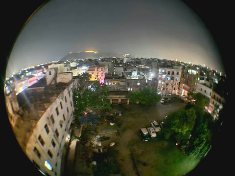       Fisheye night view of a densely packed cityscape with glowing lights and distant hills.
  