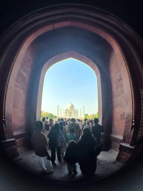       Crowds move through a grand red-sandstone arch framing a distant view of the Taj Mahal under a clear blue sky.
  
