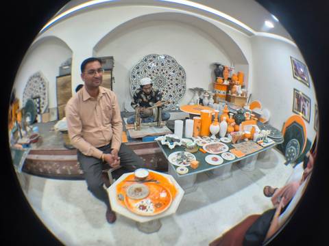       Artisan demonstrates intricate inlay work on marble plates inside a small craft workshop while a visitor observes.
  