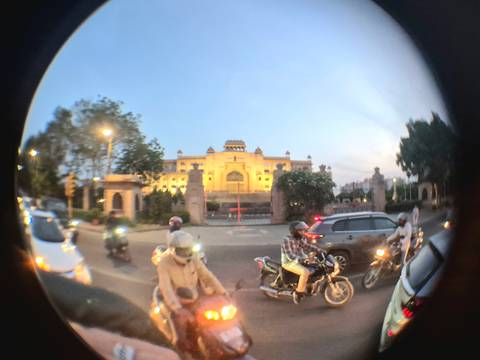       Busy evening traffic with motorbikes passing a lit heritage building, shot through a fisheye lens.
  