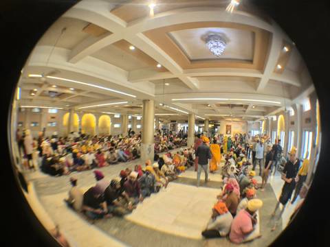       Large congregation seated on the floor of a bright hall, likely part of a communal meal at a Sikh gurdwara.
  