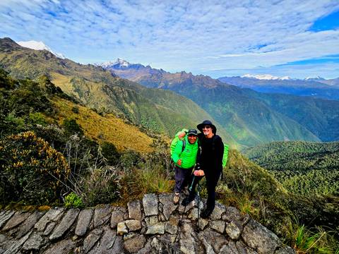       Two trekkers in bright gear smiling on a high ridge overlooking vast green ranges
  