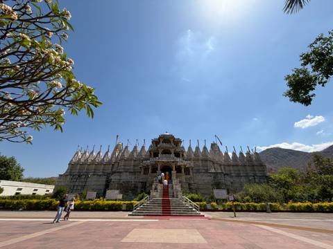       Ornate marble Jain temple with visitors ascending a red-carpeted stairway under a bright blue sky
  