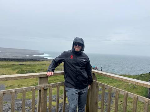       Man in a hooded jacket posing on a wooden viewing platform above windy sea cliffs under an overcast sky.
  