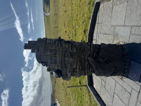       Vertical carved wooden sculpture marking the Cliffs of Moher Experience set against grassy hills and blue sky.
  