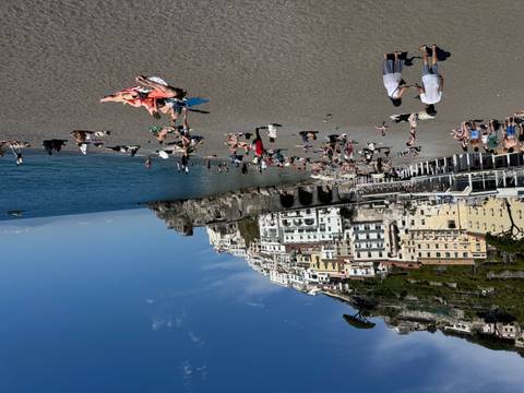       Busy beach scene in Amalfi with sunbathers and swimmers below pastel cliffside buildings and a bright blue sea.
  