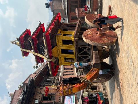       Large decorated wooden chariot on massive wheels surrounded by festival goers in a historic square.
  