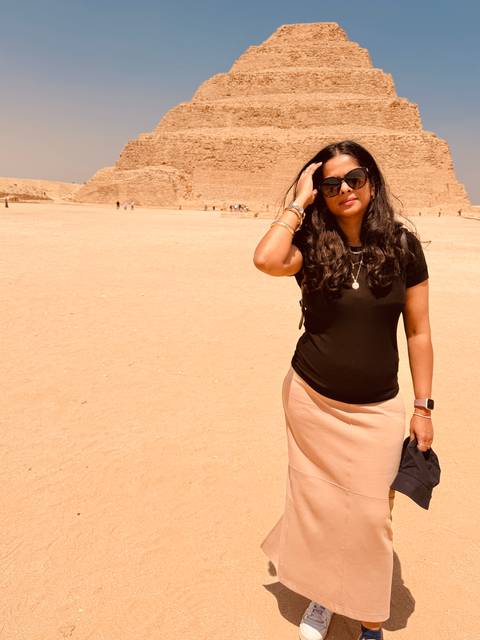       Woman in sandy foreground of pyramid complex shielding eyes from sun
  