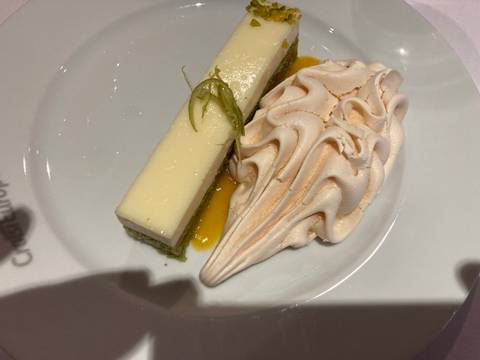       Slice of creamy cheesecake accompanied by lime zest and a large swirl of meringue on a white plate.
  