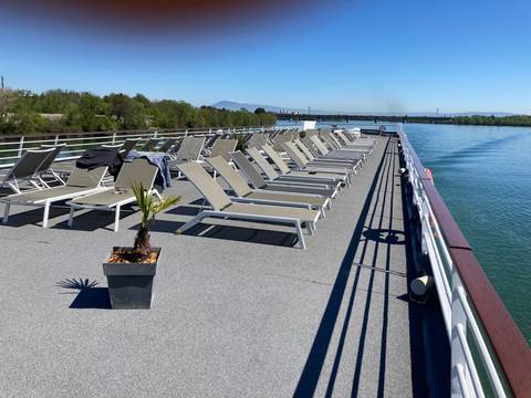       Sunny top deck of a river cruise ship lined with empty lounge chairs and calm blue water on both sides.
  