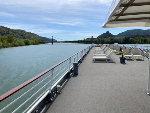       River cruise deck view toward a small suspension bridge and green hills under a bright blue sky.
  