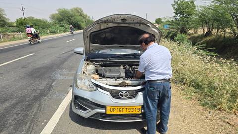       Driver inspects the engine of a silver taxi stopped on a rural highway while motorbikes pass by.
  