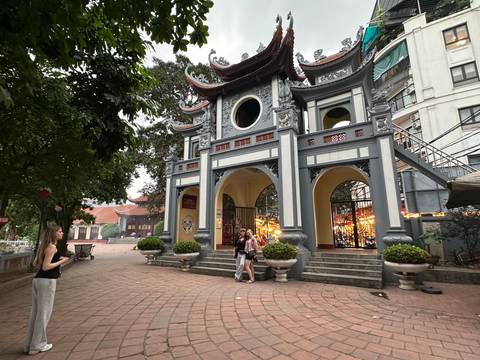       Historic pagoda-style entrance gate with ornate roof carvings, flanked by trees and visitors taking photos.
  