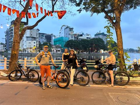       Group of cyclists posing with bikes beside an urban lake at dusk with city buildings in the background.
  