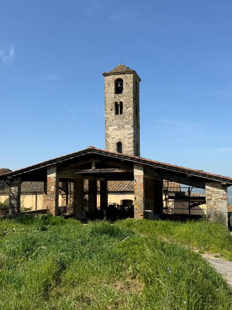       Stone bell tower of a rustic church rising above tiled roofs against a clear sky in rural Tuscany.
  