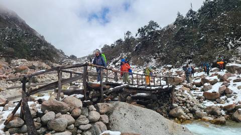       Hikers crossing a rustic wooden bridge over a snowy mountain stream
  