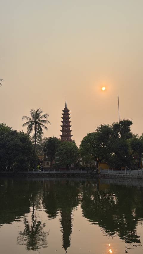       Tran Quoc Pagoda rises among trees beside West Lake under a hazy sunset sky in Hanoi.
  