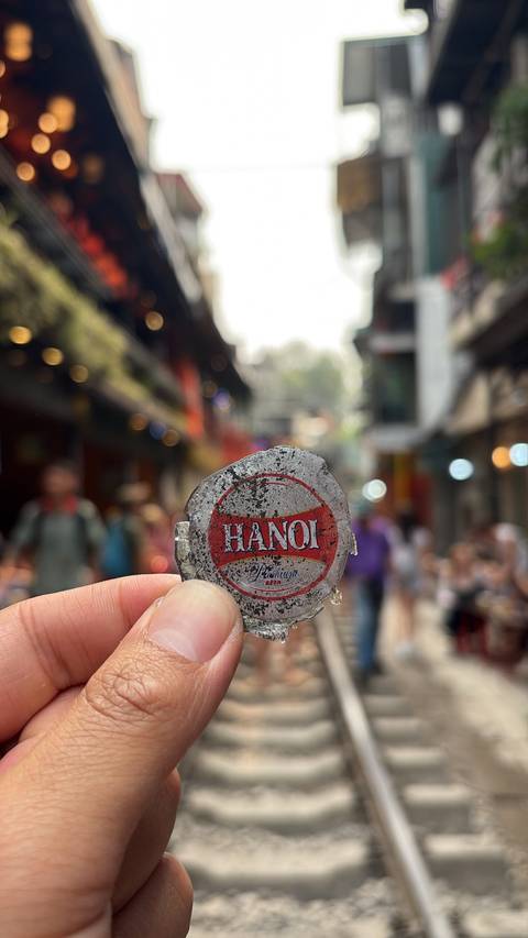       Close-up of a Hanoi beer bottlecap held up against a bustling narrow street with colorful shopfronts and blurred pedestrians in the background.
  