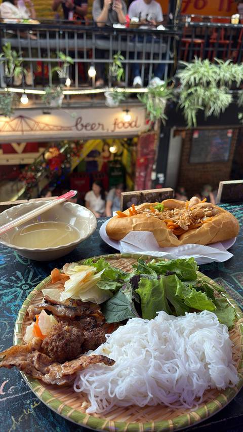       Vietnamese lunch setting with a steaming bowl of broth, a freshly made banh-mi sandwich, and salad plates on a graffiti-covered counter inside a lively eatery.
  