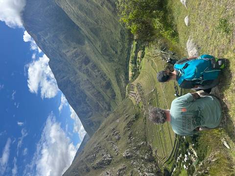       Pair of trekkers sit on a ridge overlooking ancient agricultural terraces and a verdant valley under a brilliant blue sky.
  