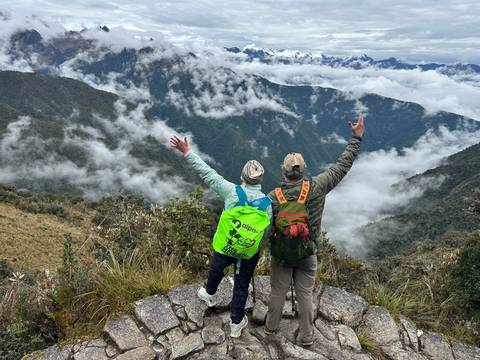       Two hikers celebrate atop a cliff surrounded by clouds and lush Andean ridges, arms raised in triumph.
  