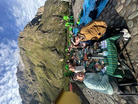       Long outdoor breakfast table set beside towering Andean peaks where trekkers enjoy an early meal at camp.
  