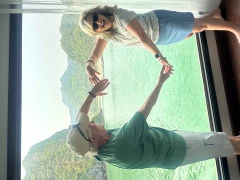       Couple forms a heart shape with their arms in front of a panoramic window overlooking green waters and karst peaks.
  