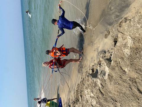       Parasailing participants wearing life vests gather at the shoreline as a guide pulls in the parachute lines from the sea.
  