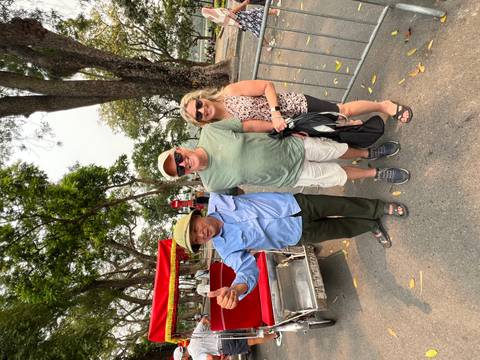       Couple poses with a cyclo driver who gestures enthusiastically on a tree-lined Hanoi street.
  
