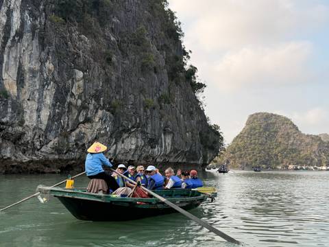       Rowboat filled with tourists glides between towering limestone cliffs on calm emerald water in Halong Bay.
  
