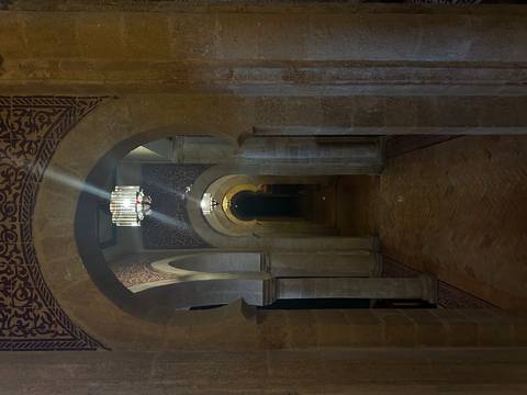       Dimly lit stone corridor of a Moroccan mosque or palace with repeating arches and hanging chandeliers.
  