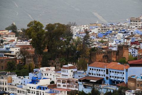       Panoramic view over Chefchaouen’s blue and white buildings nestled among green hills.
  