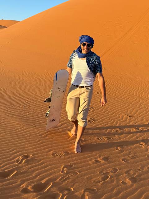      Adventurer carrying a sandboard barefoot across rippled orange dunes of the Sahara at sunset.
  