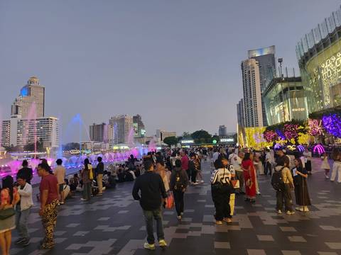       Crowds gather along a riverside mall with colorful fountains and city skyline lights at dusk in Bangkok.
  