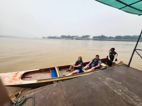       Two tourists seated in a long-tail boat with driver on a wide muddy river
  