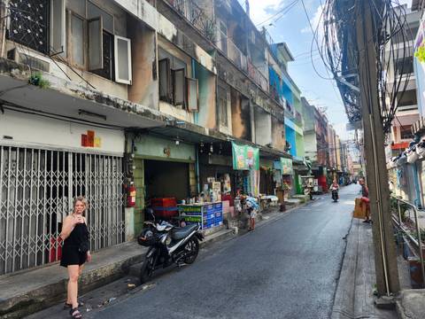       Narrow urban lane with older buildings, small shops and a tourist sipping a drink
  
