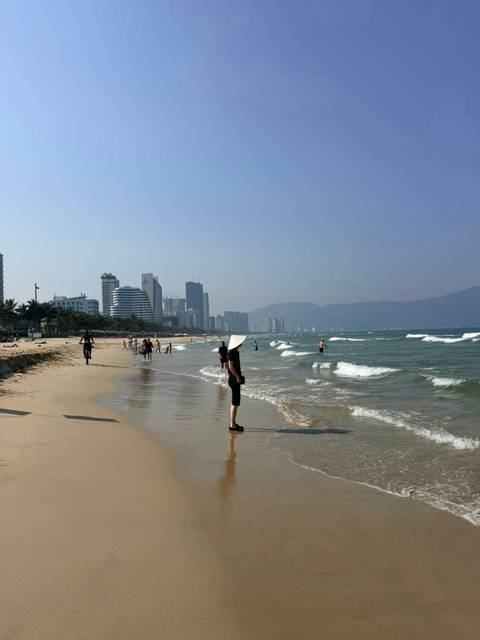       Wide sandy beach at Da Nang with city skyline and gentle waves, woman in conical hat in foreground
  
