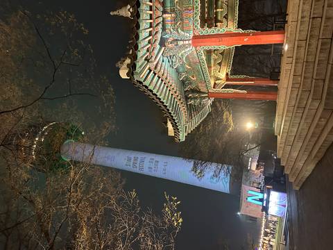       Night scene of Namsan Seoul Tower lit for spring festival beside a traditional pavilion and bare tree branches.
  