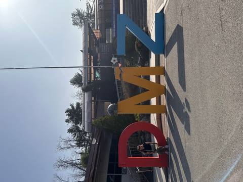       Large coloured DMZ letters in front of visitor centre with single tourist seated beside the sign on sunny day.
  