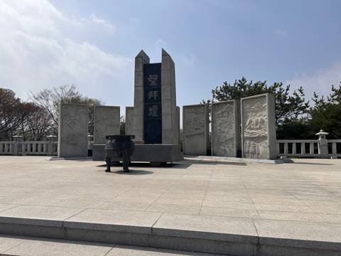       Outdoor stone memorial monument flanked by carved panels under partly cloudy sky.
  