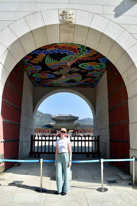       Visitor framed under ornate painted palace archway looking toward main throne hall with mountains behind.
  