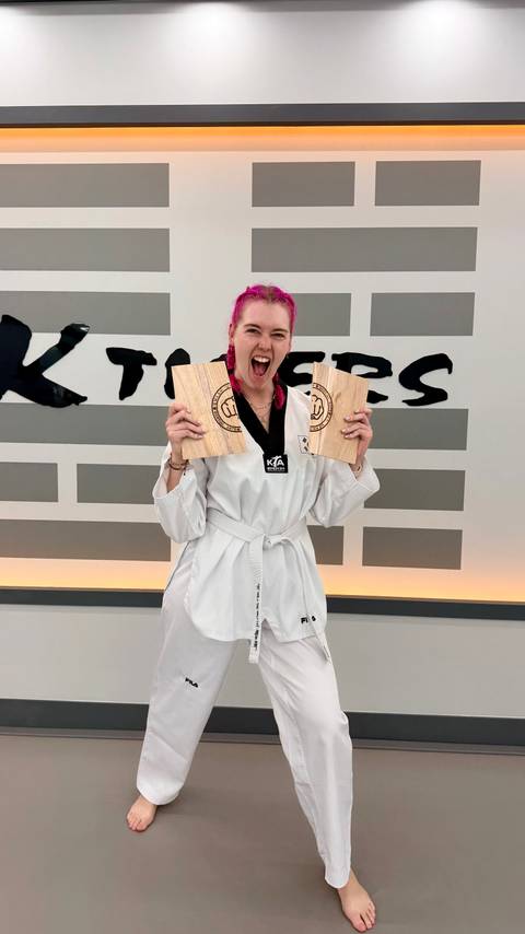       Excited woman in taekwondo uniform proudly holding two broken boards at martial arts studio.
  