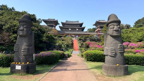       Grand entrance path flanked by two stone dolhareubang statues leading to a multi-tiered Korean temple surrounded by colorful azaleas.
  