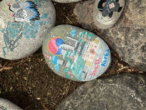       Painted souvenir stone depicting South Korea landmarks lying among rocks.
  