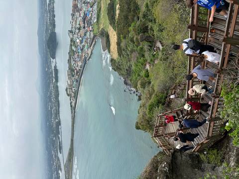       Wooden boardwalk filled with hikers descending from a volcanic peak overlooking coastal town and bay.
  