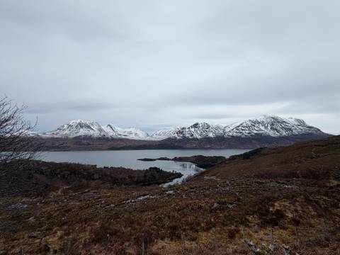       Wide view of snow-covered Torridon peaks across a still loch under grey sky.
  