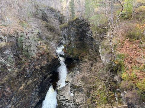       Deep rocky gorge with narrow river cutting through layered stone walls and sparse trees.
  