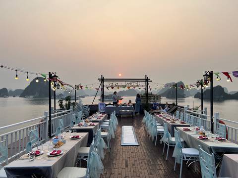       Elegant outdoor dining setup on a boat deck cruising through limestone islets under a hazy sunset sky
  