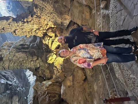       Two visitors posing inside an illuminated limestone cave with dramatic stalactite formations
  